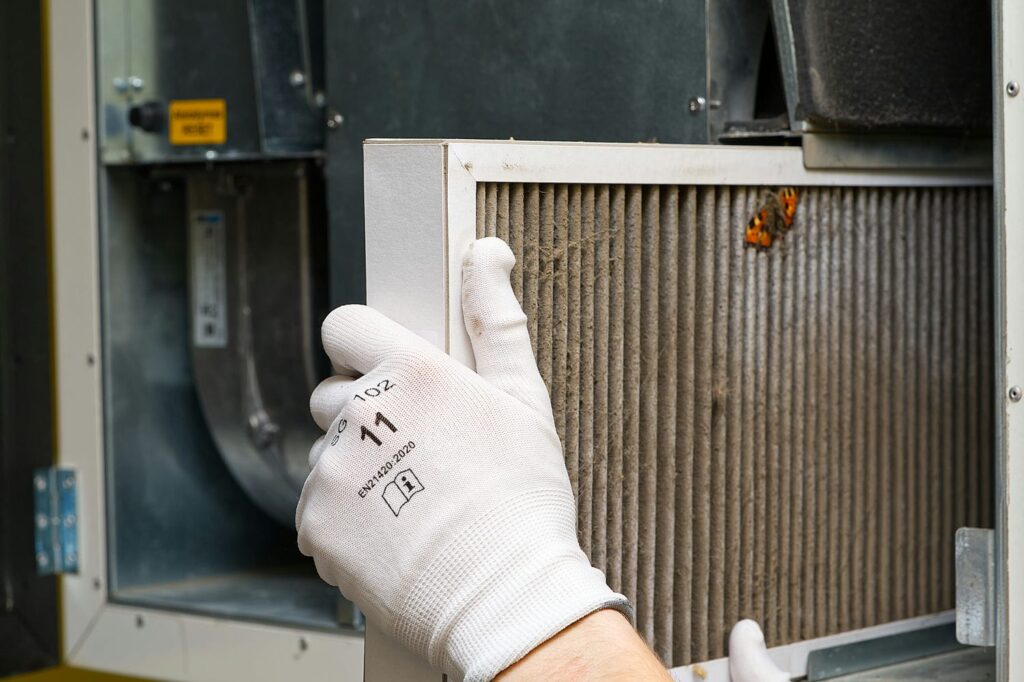 Homeowner sliding a dusty pleated filter out of the return air slot beside a gas furnace and duct mounted humidifier in a basement
